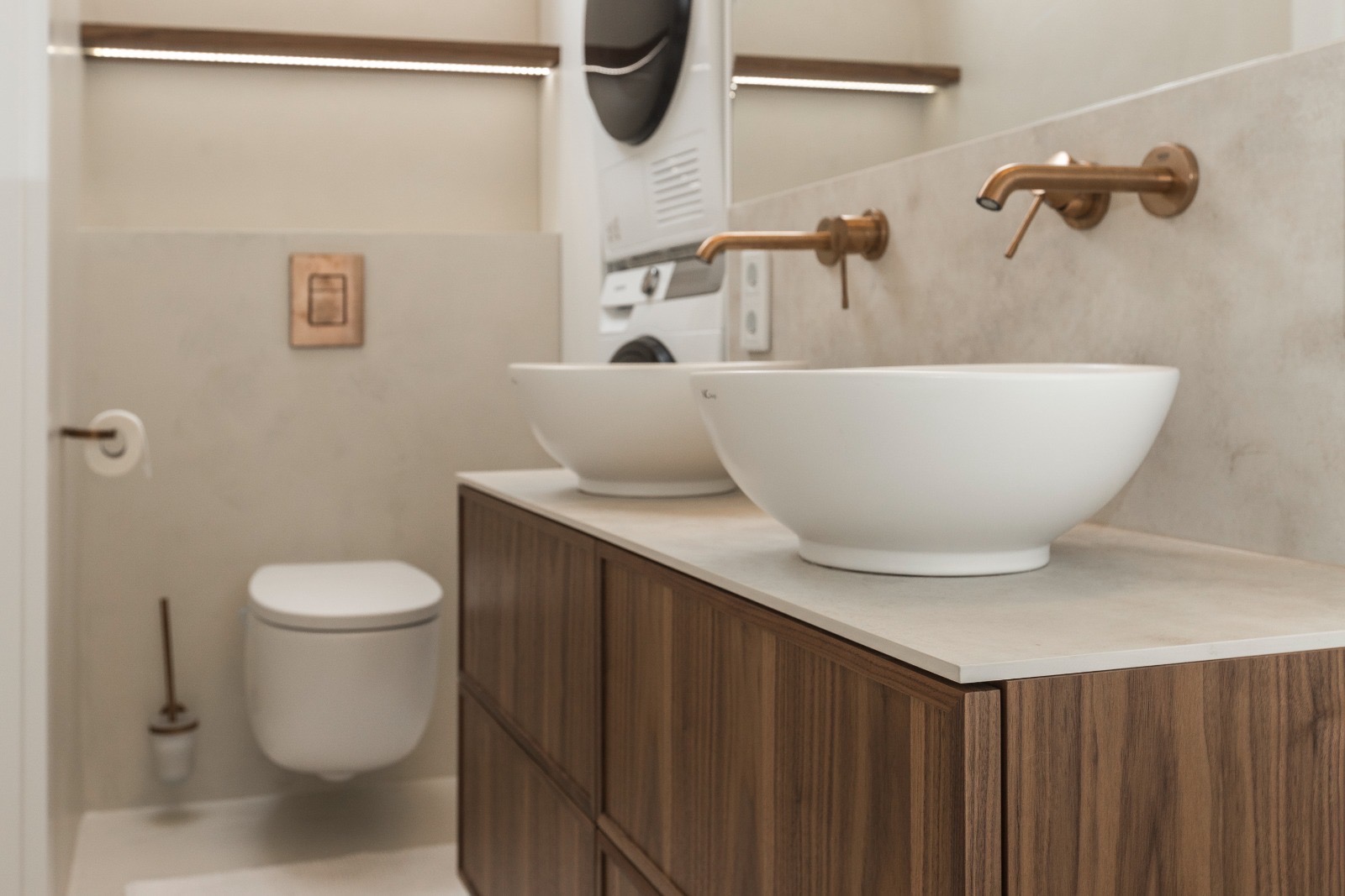 Nowoczesna łazienka z dwoma białymi umywalkami na drewnianej szafce, armaturą w kolorze miedzi i podświetlanymi półkami. W tle pralka i suszarka.