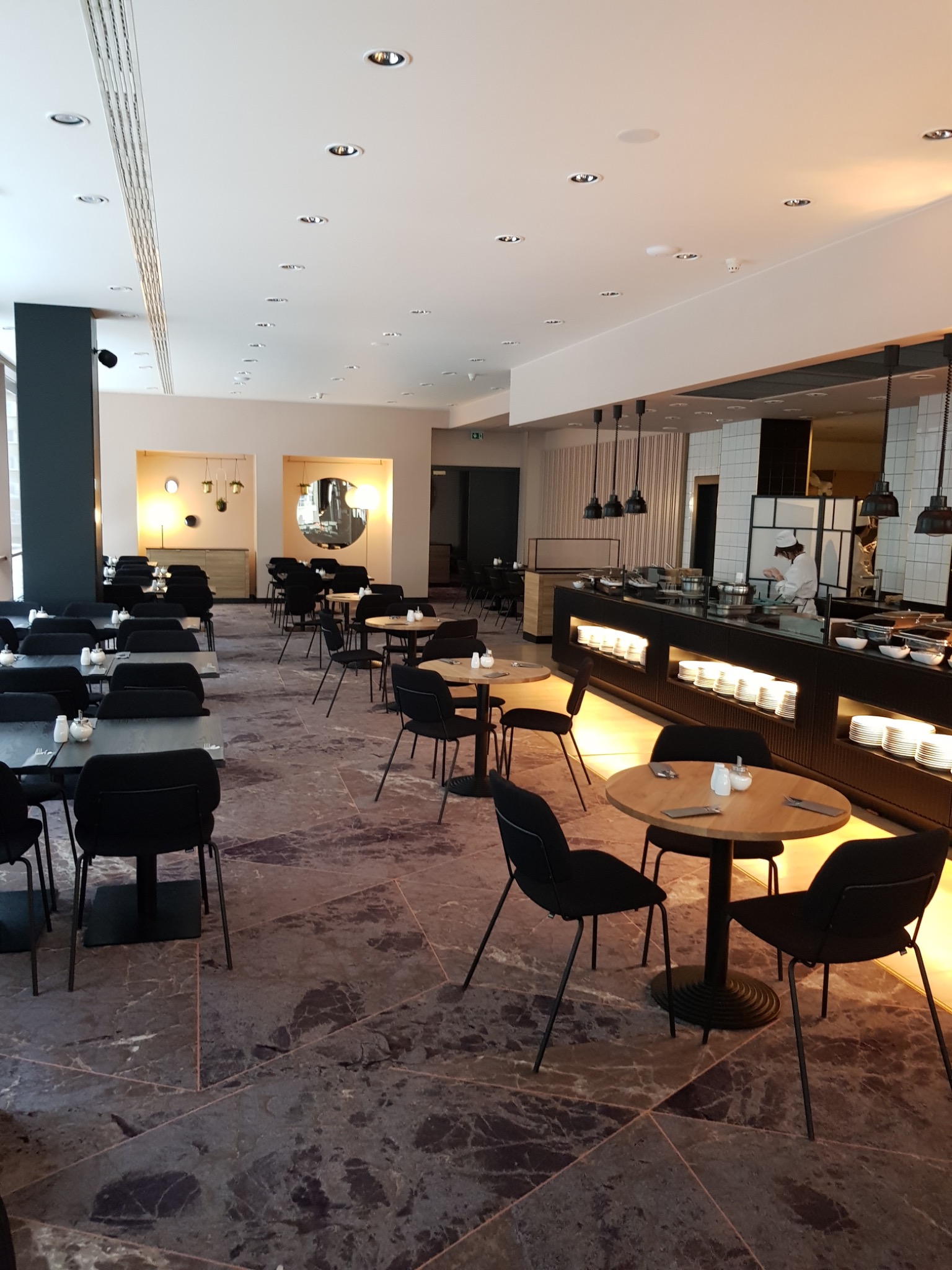 Elegancka restauracja z marmurową podłogą, okrągłymi stolikami i czarnymi krzesłami. W tle widoczny bufet z kucharzem przygotowującym dania.