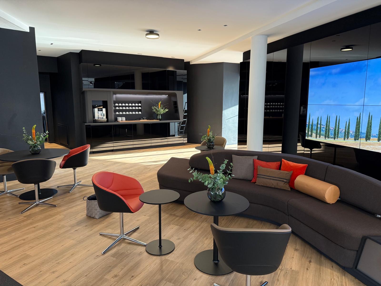 Nowoczesne wnętrze z czarną zabudową kuchenną, brązową sofą i kolorowymi krzesłami. Na ścianie duży ekran z widokiem na toskański krajobraz.
