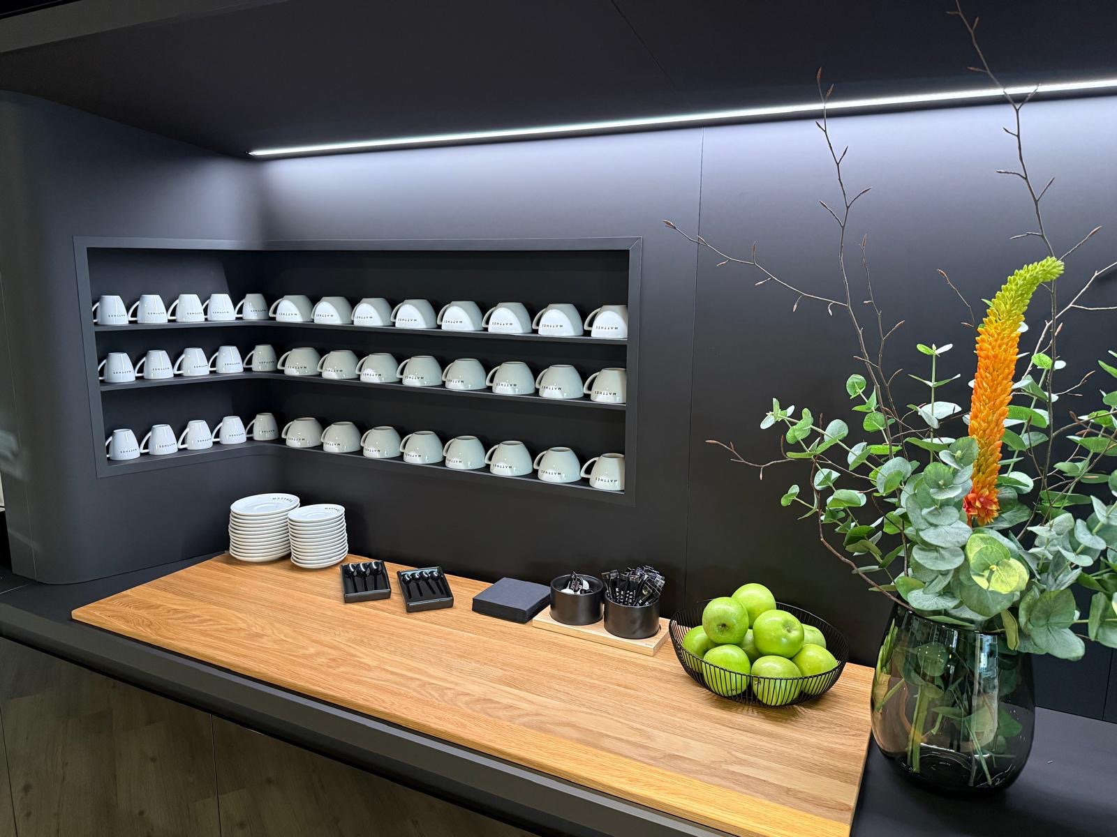 Nowoczesna zabudowa kuchenna w odcieniach szarości z drewnianym blatem, dekoracyjnymi naczyniami i owocami. Minimalistyczny design z akcentami roślinnymi.