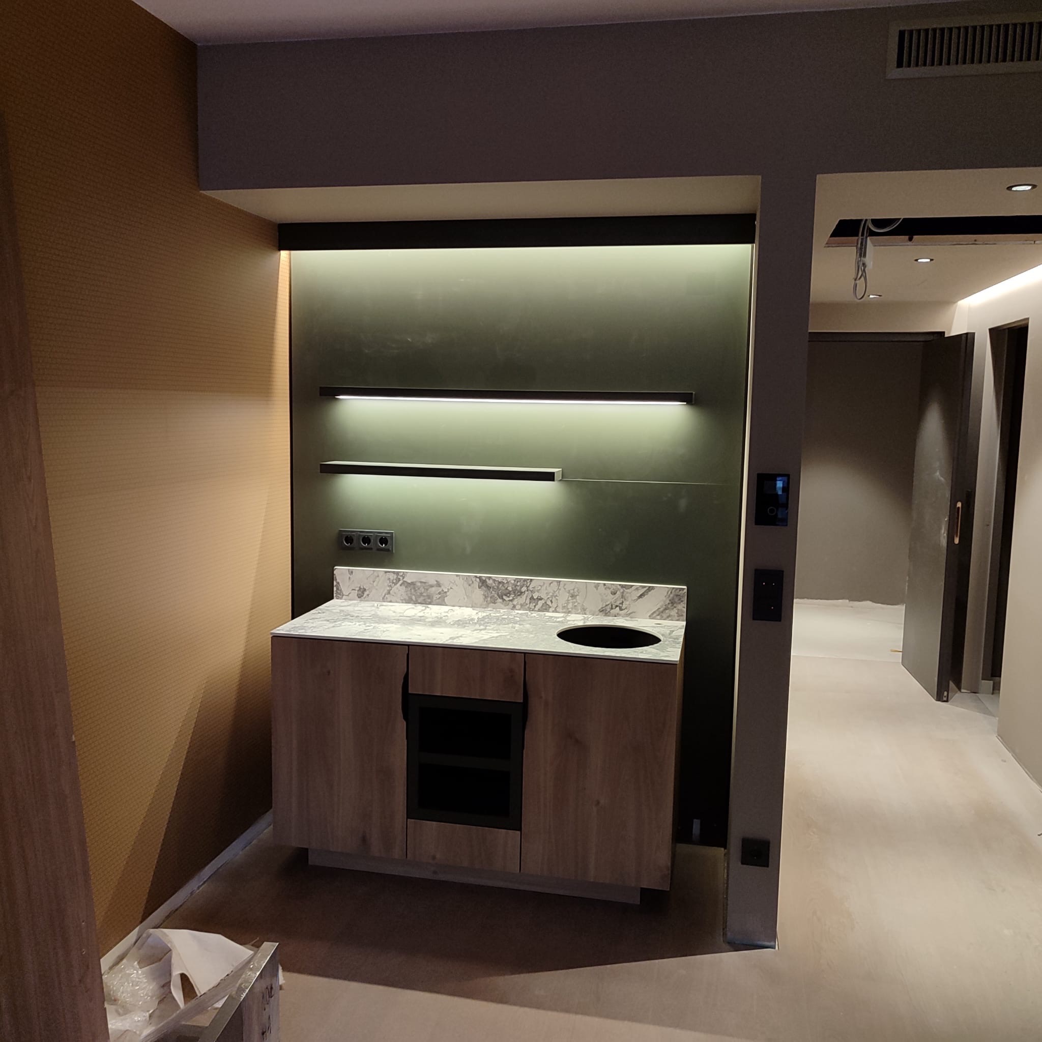 Nowoczesna zabudowa z umywalką i marmurowym blatem, oświetlona ledowymi lampami. Widoczne gniazdka elektryczne i fragment korytarza z drzwiami.