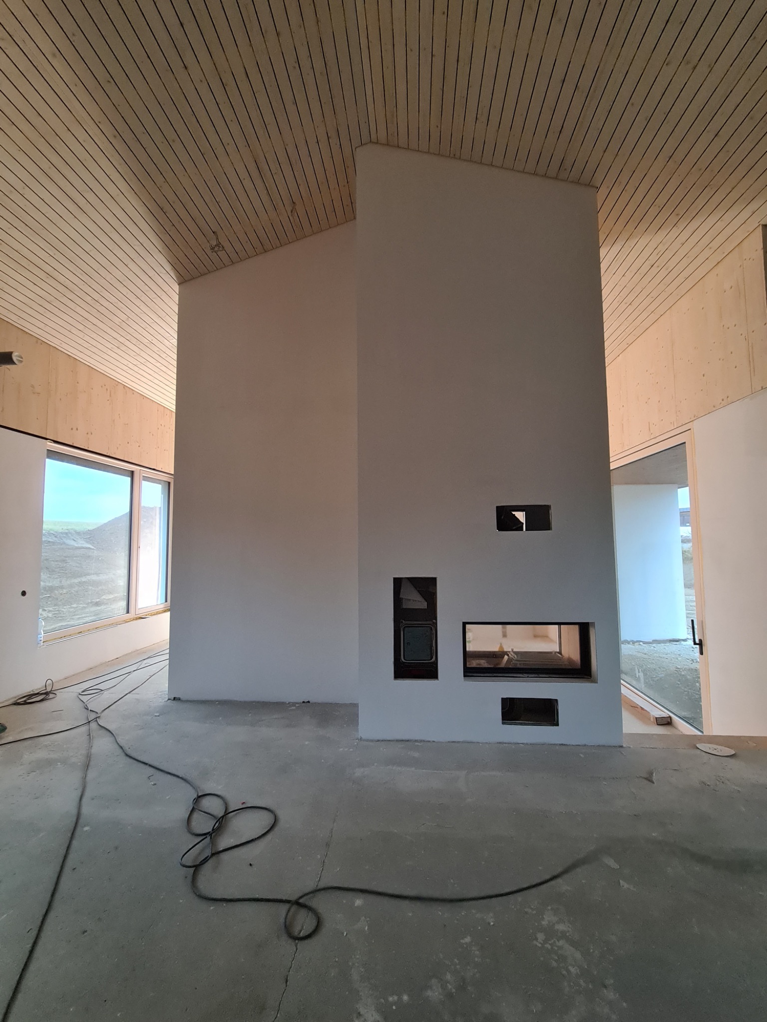 Surowe wnętrze z drewnianym sufitem i kominkiem o minimalistycznym designie. Widoczne kable na podłodze sugerują etap wykończeniowy.