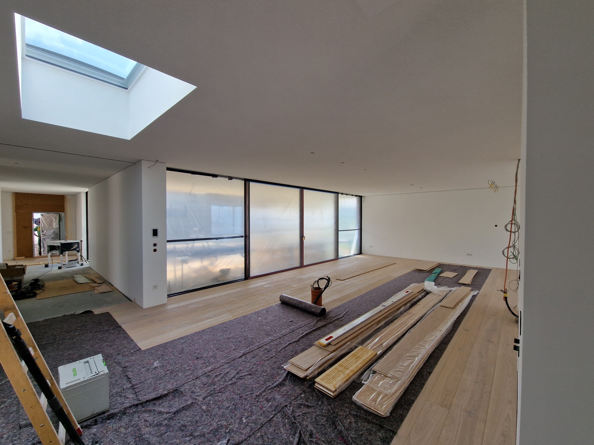 Jasne panele podłogowe w trakcie montażu w przestronnym wnętrzu z dużymi oknami i świetlikiem. Widoczne narzędzia i materiały przygotowane do dalszych prac.