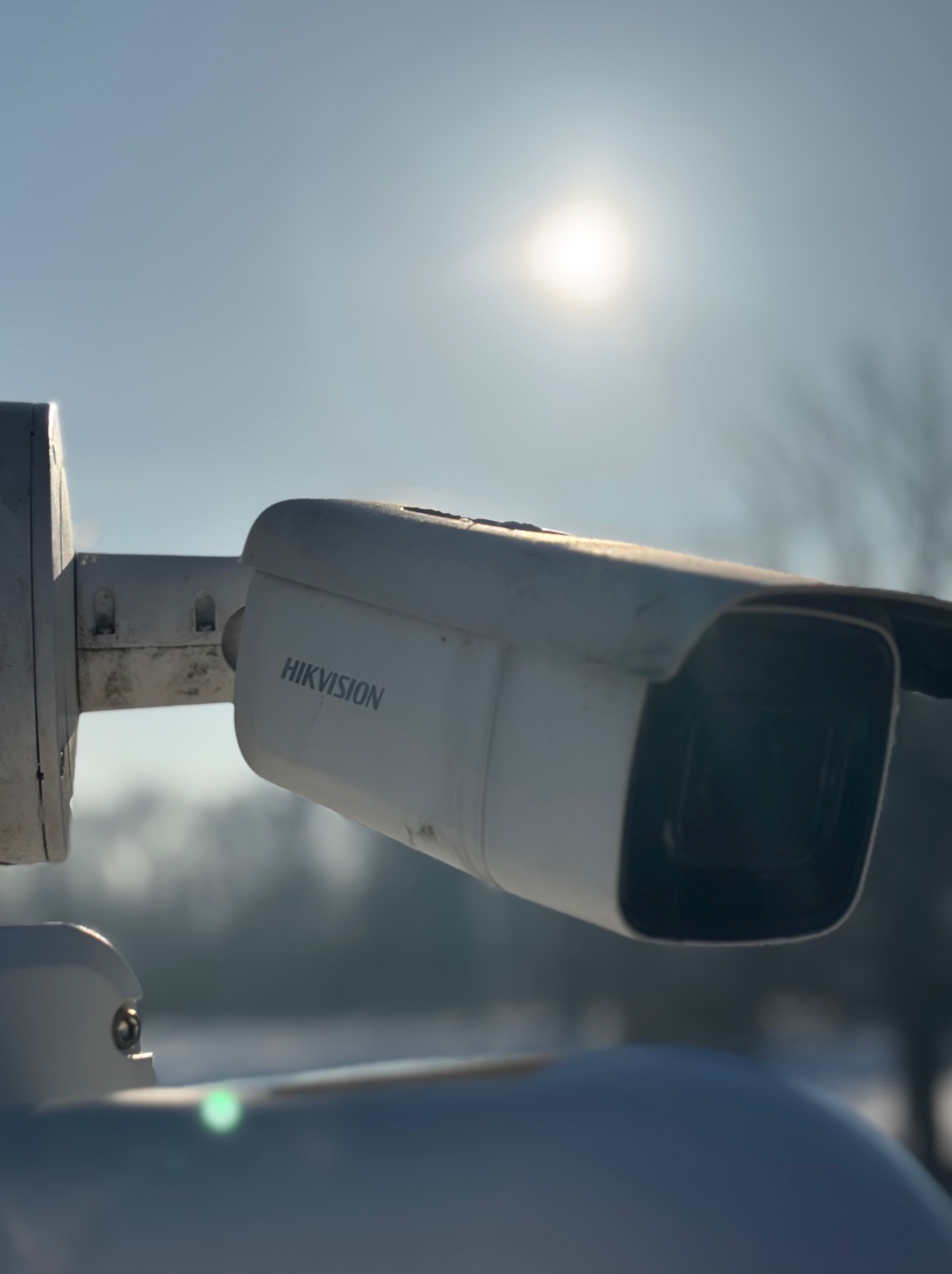 Kamera monitoringu Hikvision na zewnątrz, skierowana w górę, z widocznym słońcem w tle. Obudowa kamery lekko zabrudzona, co sugeruje jej użytkowanie na zewnątrz.