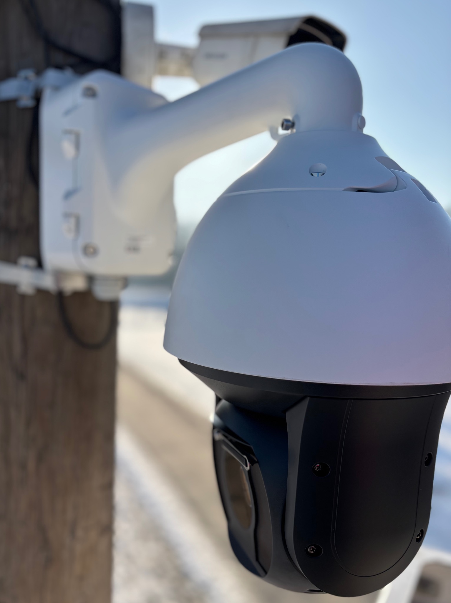 Kamera monitoringu PTZ (Pan-Tilt-Zoom) zamontowana na słupie, z widocznym uchwytem i kablem. W tle rozmyty krajobraz i błękitne niebo.