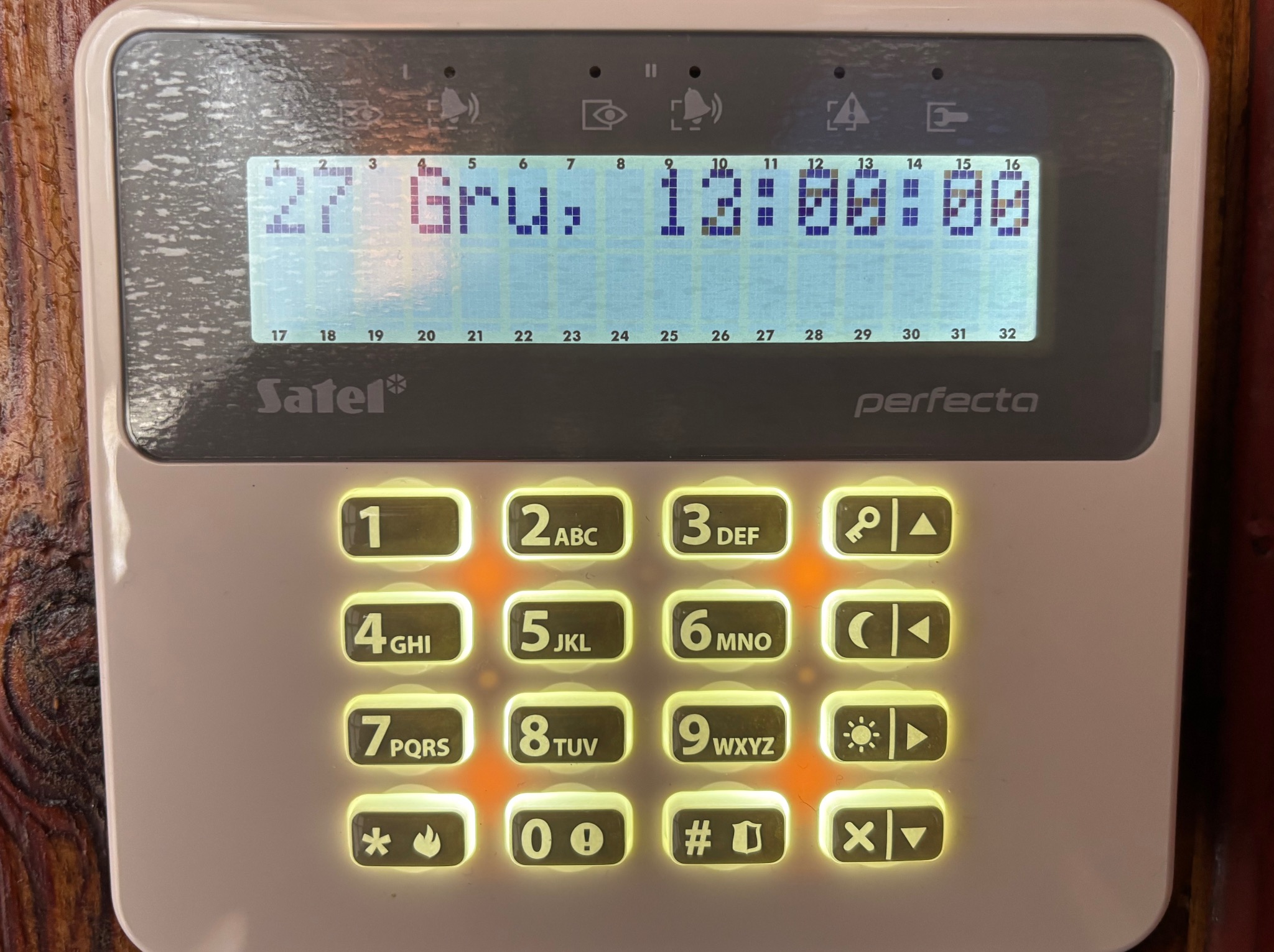 Panel sterowania alarmu Satel Perfecta z podświetlanymi przyciskami i wyświetlaczem LCD pokazującym datę i godzinę. Widoczny fragment drewnianej ściany w tle.