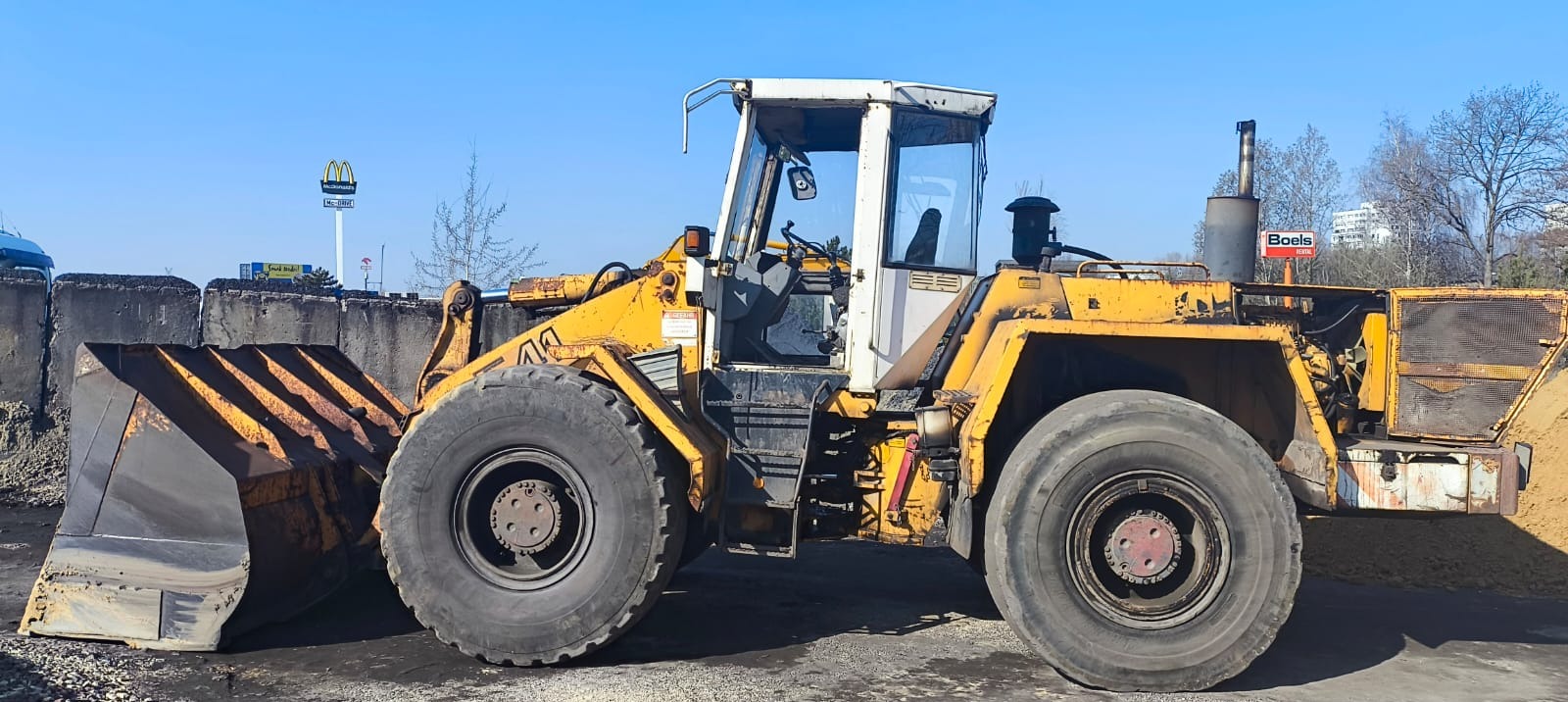 Żółta ładowarka kołowa na placu budowy, widoczny duży kubeł i opony. W tle betonowe bloki, znak McDonald's i budynki. Sprzęt budowlany w trakcie pracy.