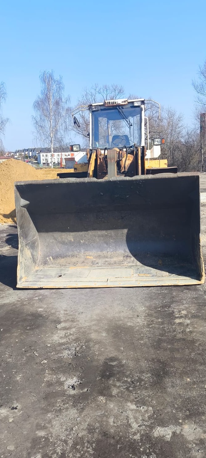 Żółta ładowarka kołowa z dużą łyżką na pierwszym planie, w tle budynki mieszkalne i hałda ziemi, błękitne niebo.