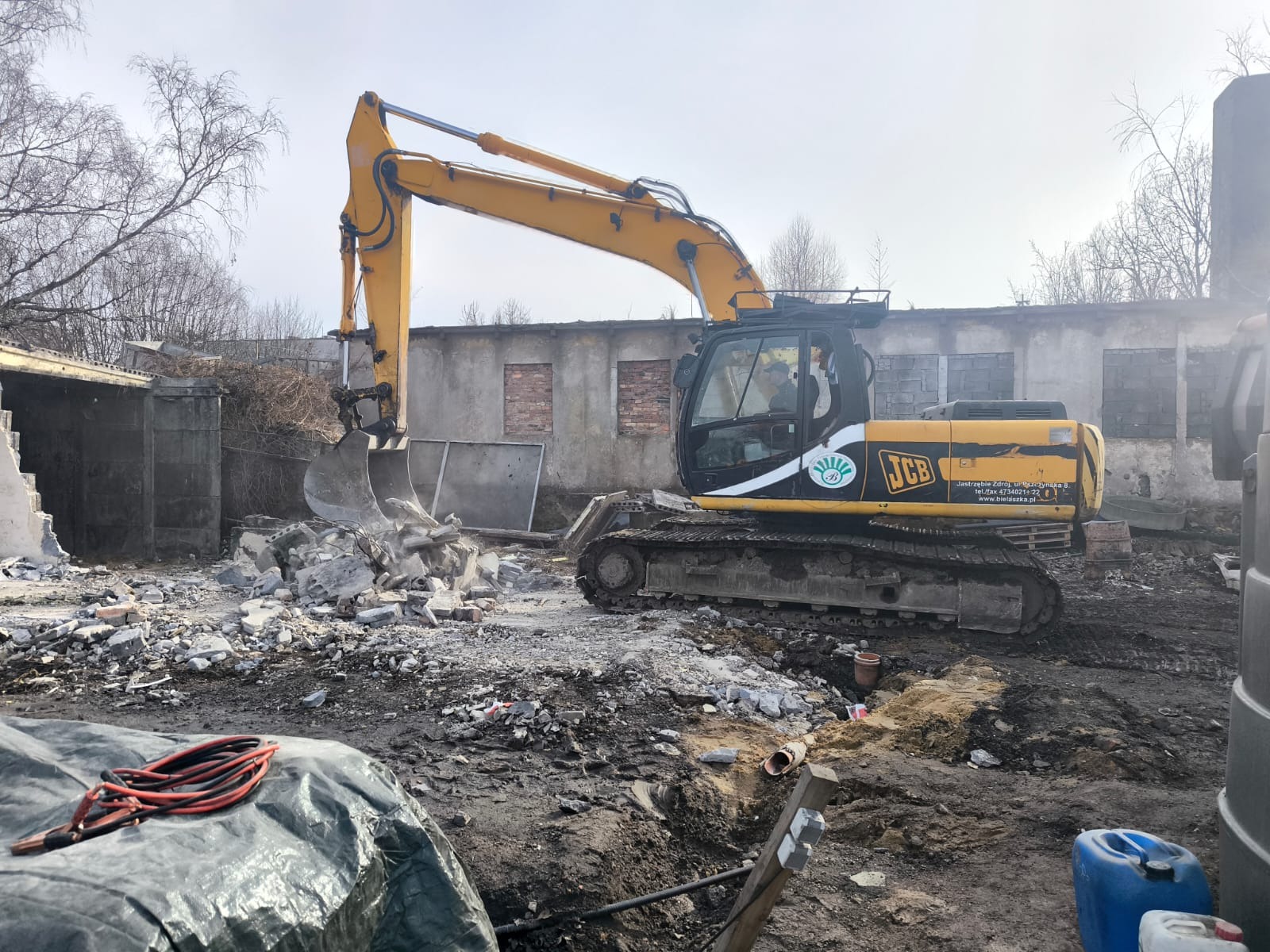 Żółta koparka JCB w trakcie rozbiórki budynku, gruz w łyżce, widoczne fragmenty zburzonej konstrukcji i ślady pracy w ziemi. Praca w toku, pochmurny dzień.