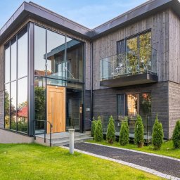 AK INVEST-BUD - Nowoczesny dom z drewnianą elewacją i dużymi przeszkleniami. Widoczny balkon z szklaną balustradą, zadbany trawnik i ścieżka prowadząca do wejścia.