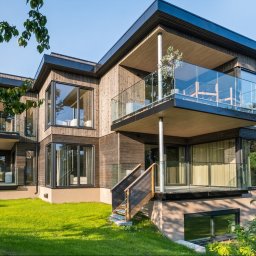 AK INVEST-BUD - Nowoczesny dom z drewnianą elewacją i balkonami z szklanymi balustradami, otoczony zielonym trawnikiem w słoneczny dzień. Widoczne duże okna i minimalistyczny design.