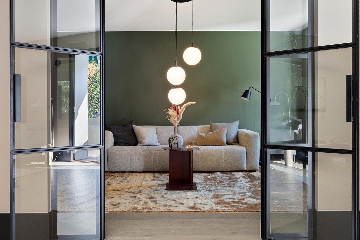 Nowoczesny salon z otwartymi, przeszklonymi drzwiami w czarnych ramach. Wnętrze z beżową sofą, dywanem w odcieniach brązu i zieloną ścianą. Minimalistyczne lampy wiszące.
