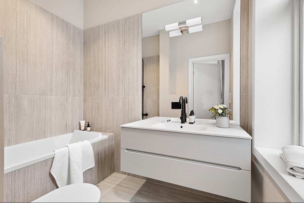 Nowoczesna łazienka z wanną, umywalką w zabudowie i dużym lustrem. Ściany wyłożone płytkami imitującymi drewno. Minimalistyczny design, czarna armatura, kwiaty w wazonie.