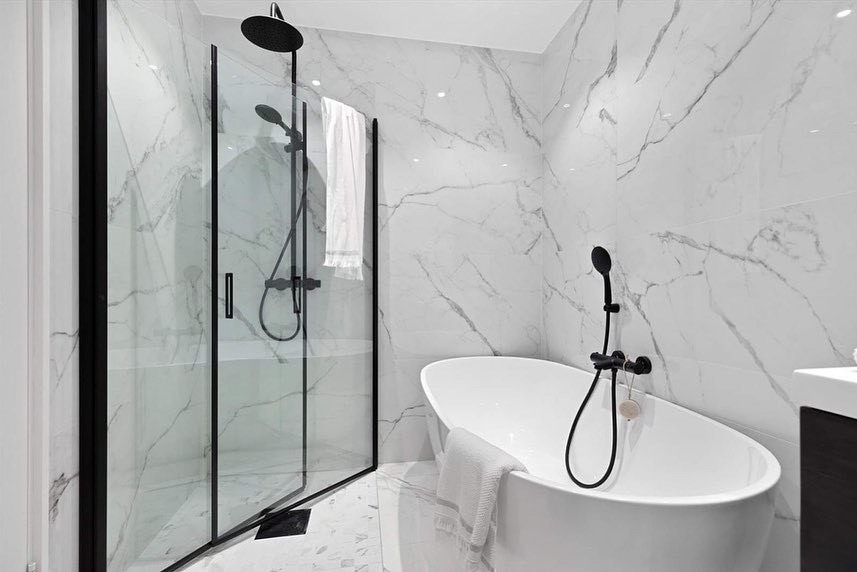 Nowoczesna łazienka z kabiną prysznicową w czarnej ramie i wolnostojącą wanną na tle białych marmurowych płytek. Minimalistyczny design z czarną armaturą.