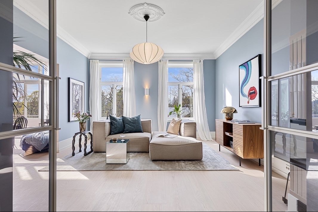 Jasne wnętrze salonu z błękitnymi ścianami, beżową sofą modułową, drewnianą komodą i nowoczesnym oświetleniem. Widok przez przeszklone drzwi balkonowe.