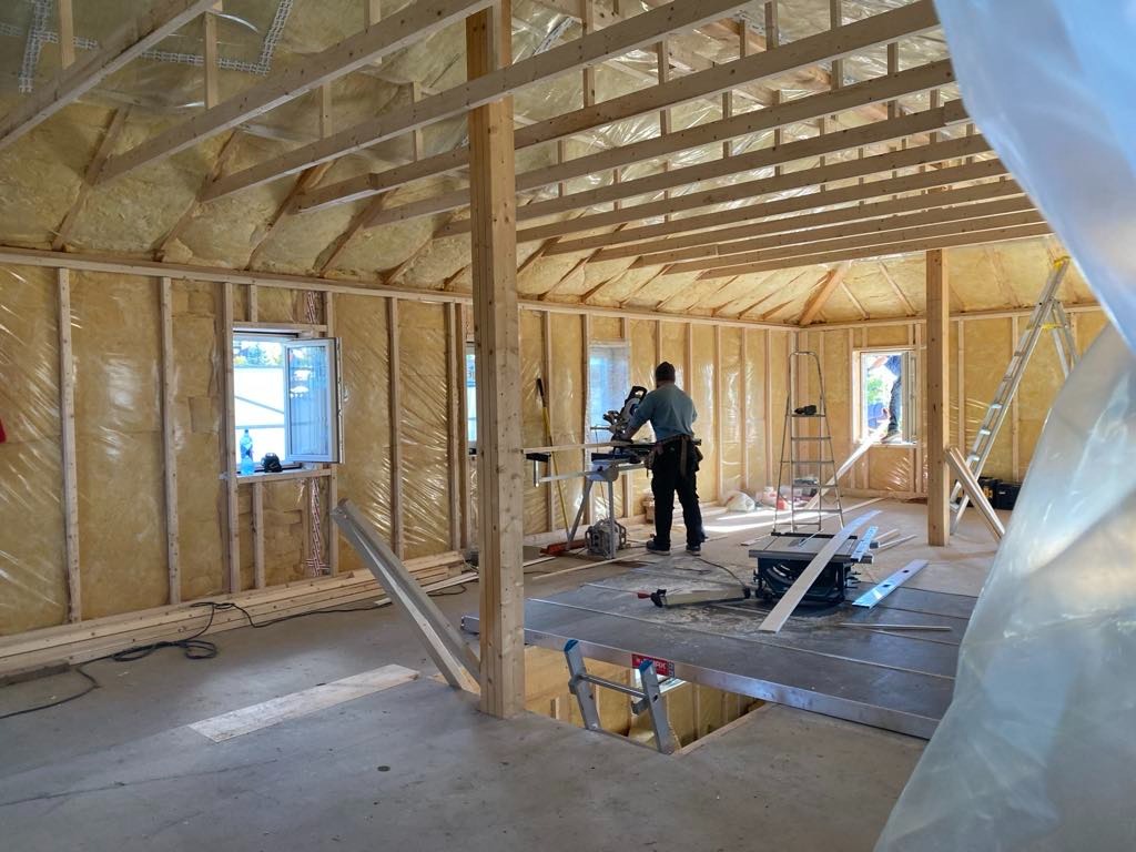 Wnętrze domu w trakcie budowy: ocieplone ściany, drewniana konstrukcja dachu, pracownik przy pile stołowej. Widoczne okna i drabiny.