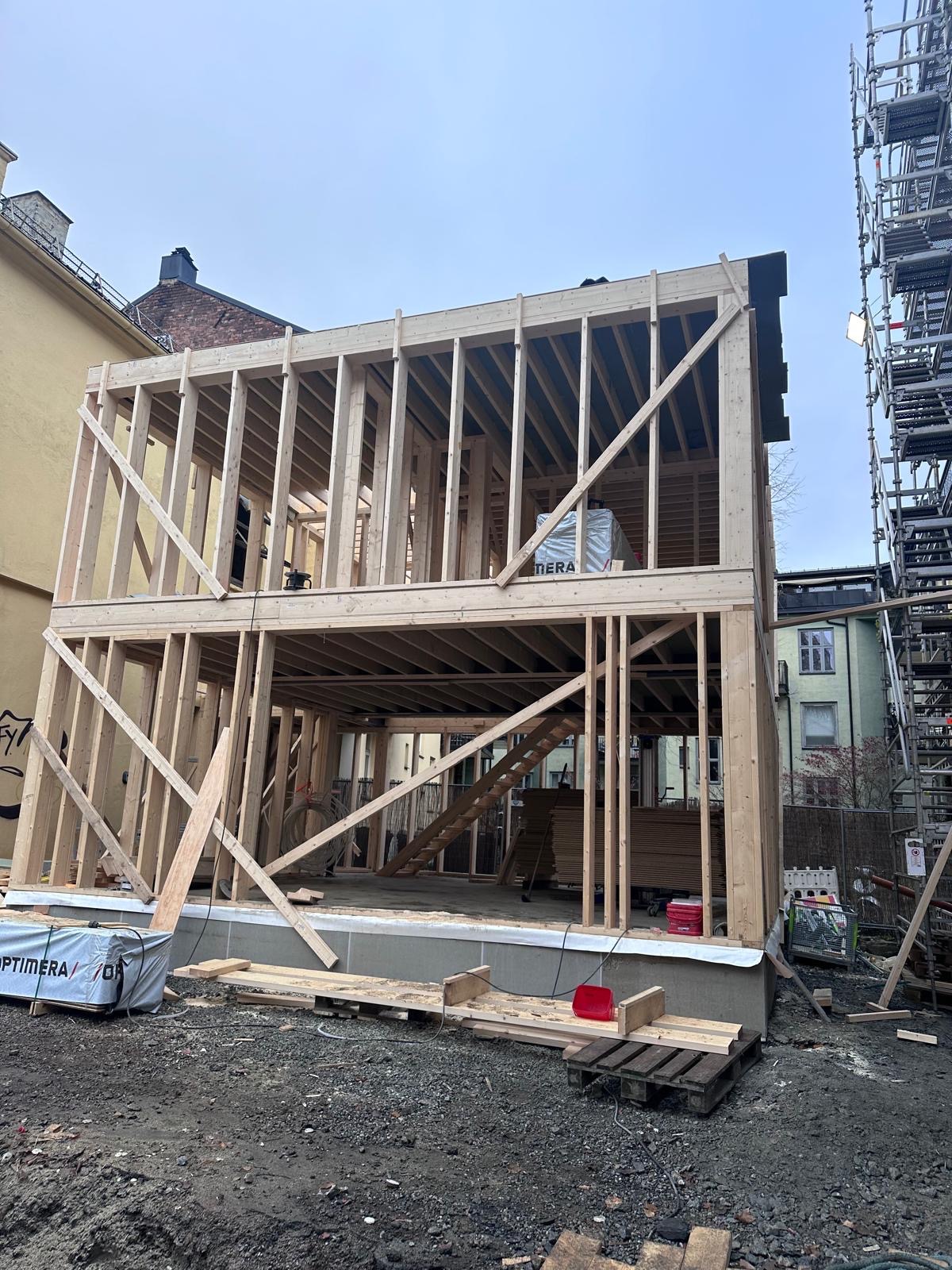 Dwukondygnacyjny dom szkieletowy w trakcie budowy, widoczne drewniane ramy konstrukcyjne, materiały budowlane i rusztowania w tle. Ujęcie z dołu, pochmurne niebo.