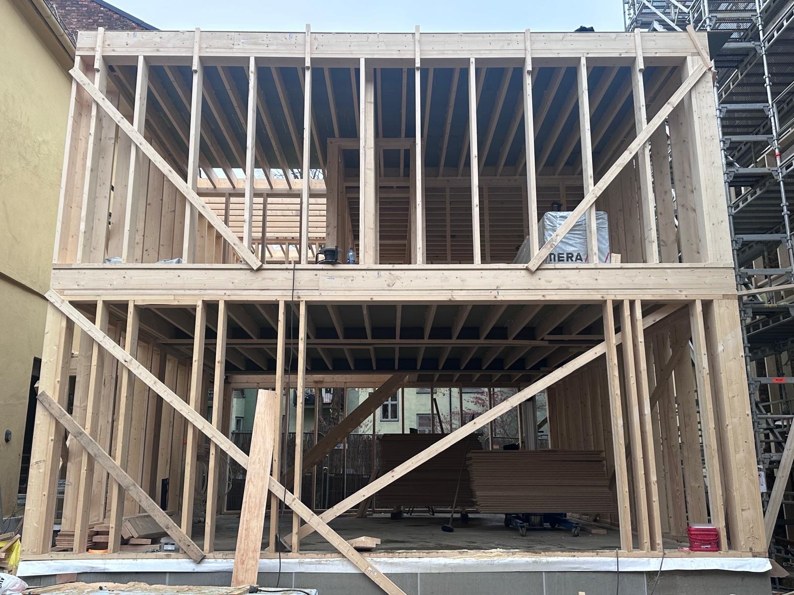 Dwukondygnacyjna konstrukcja domu szkieletowego w trakcie budowy, widoczne drewniane elementy nośne i poszycie dachu. Na drugim planie budynki mieszkalne.