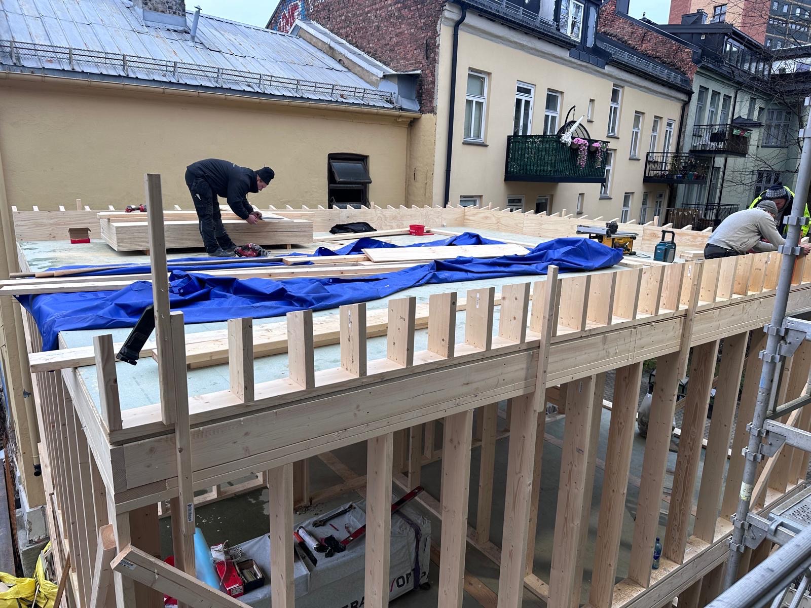 Konstrukcja szkieletowa budynku w trakcie montażu, widoczni pracownicy i narzędzia, niebieska plandeka zabezpieczająca, budynki w tle.