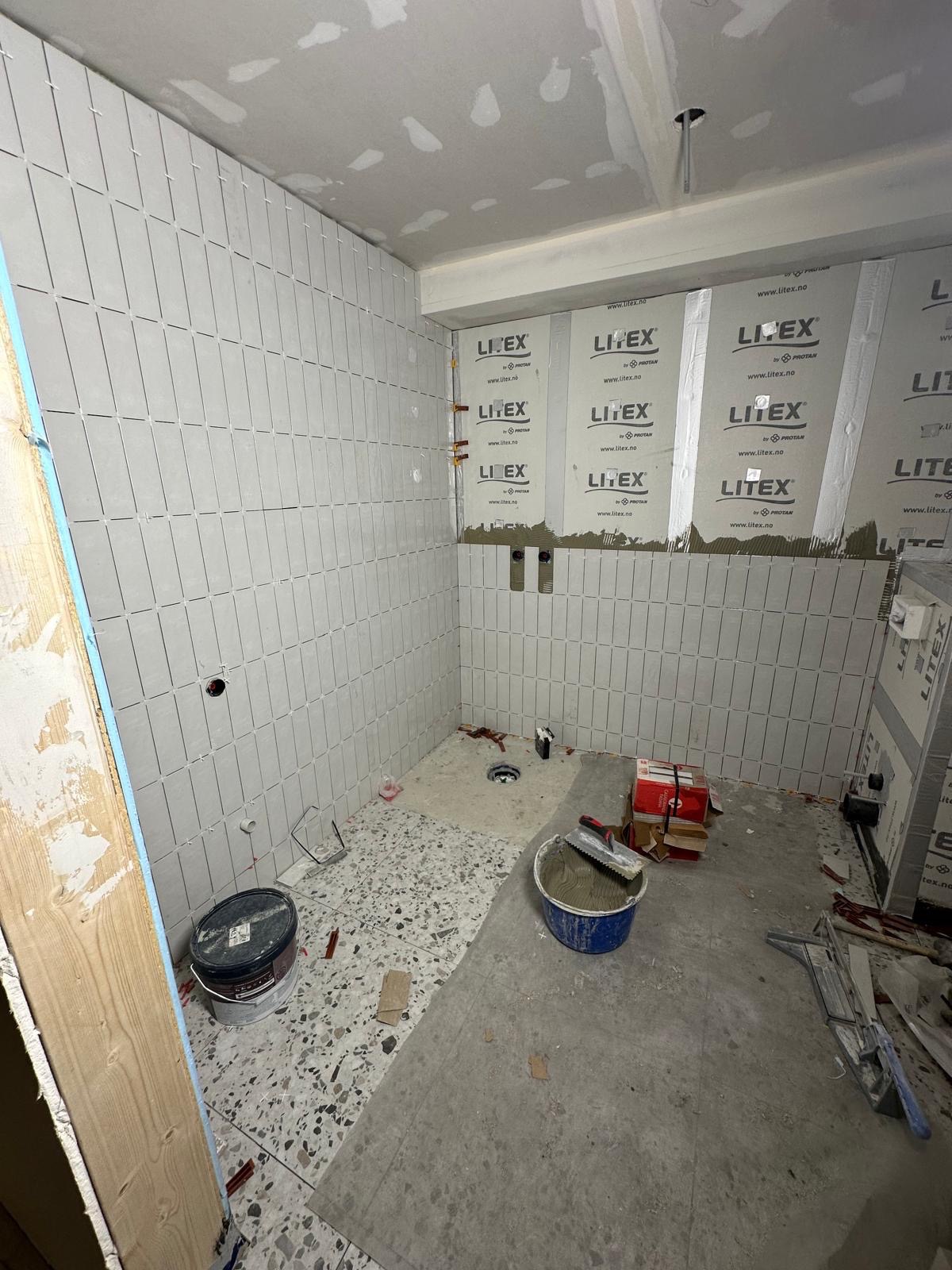 Wnętrze łazienki w trakcie remontu: ściany wyłożone szarymi, podłużnymi płytkami, częściowo zabezpieczone folią. Na podłodze widoczne narzędzia i materiały budowlane.