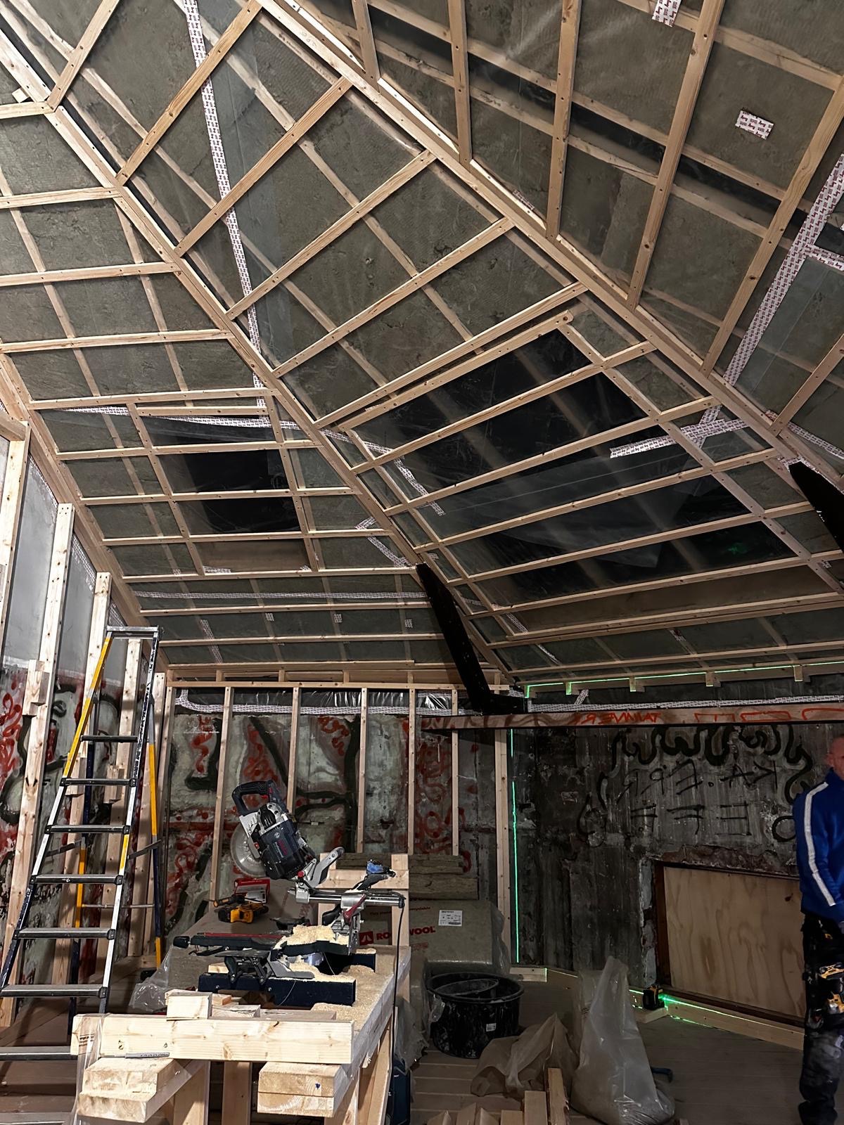 Skośny sufit z drewnianą konstrukcją i izolacją z wełny mineralnej, widoczne narzędzia i drabina, ściany z graffiti. Prace wykończeniowe w surowym wnętrzu.