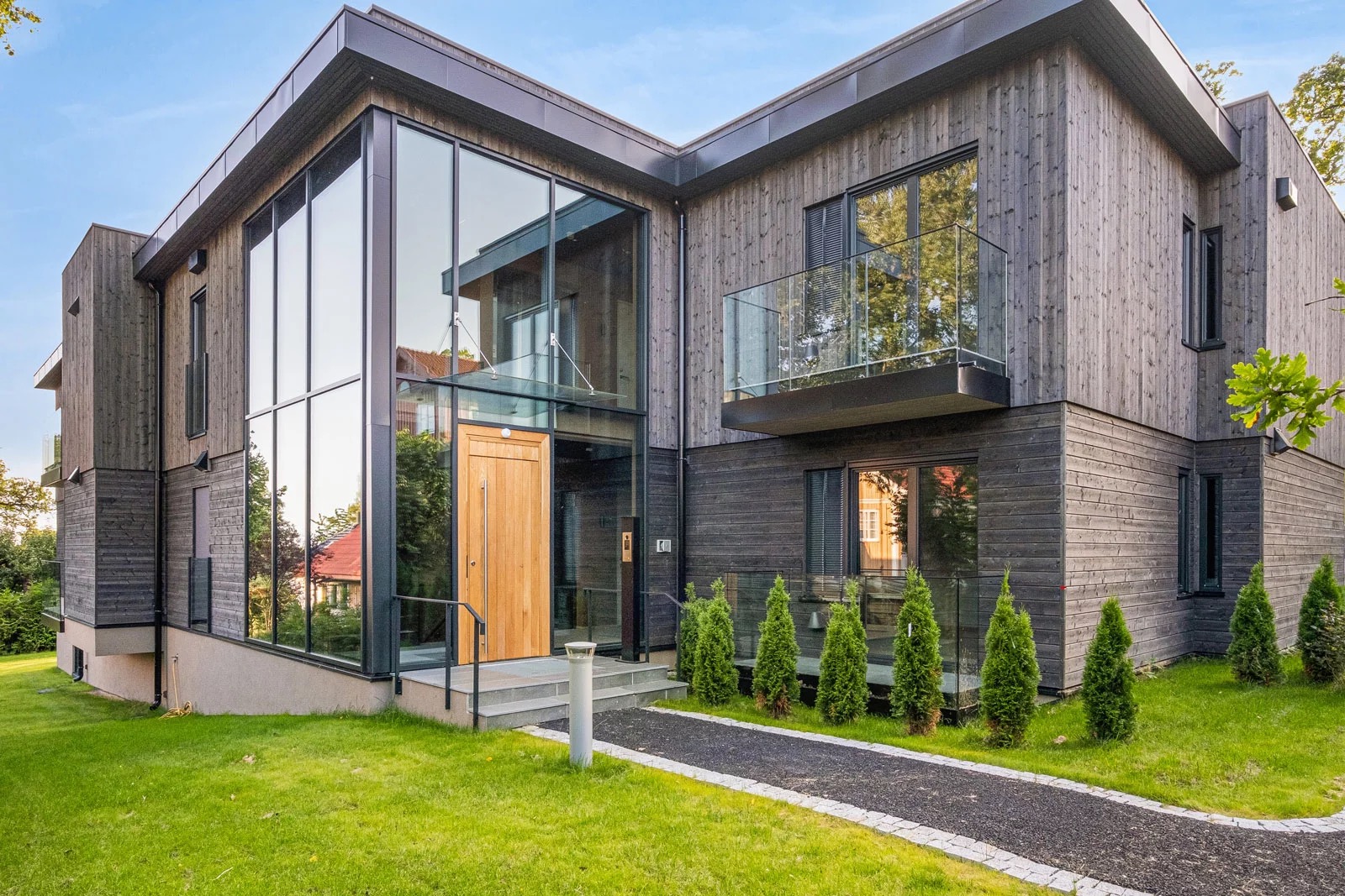 Nowoczesny dom z drewnianą elewacją i dużymi przeszkleniami. Widoczny balkon z szklaną balustradą, zadbany trawnik i ścieżka prowadząca do wejścia.