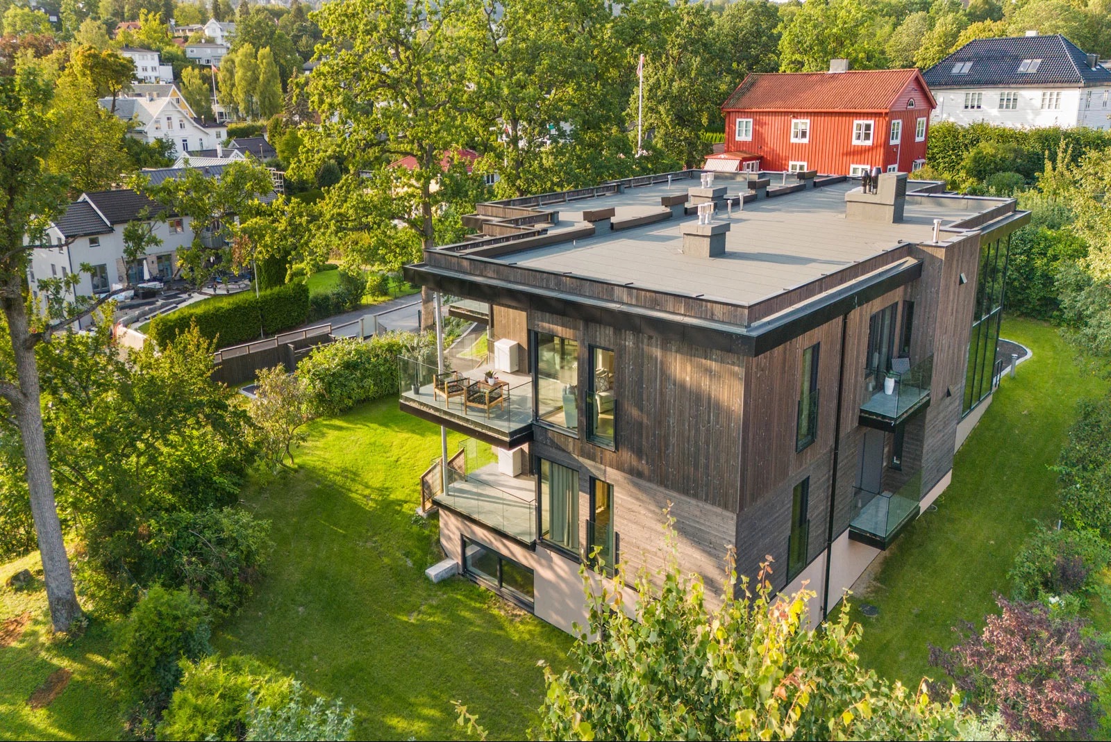 Nowoczesny dom z drewnianą elewacją i szklanymi balkonami, otoczony zielenią. Widok z góry ukazuje płaski dach i minimalistyczny design budynku.