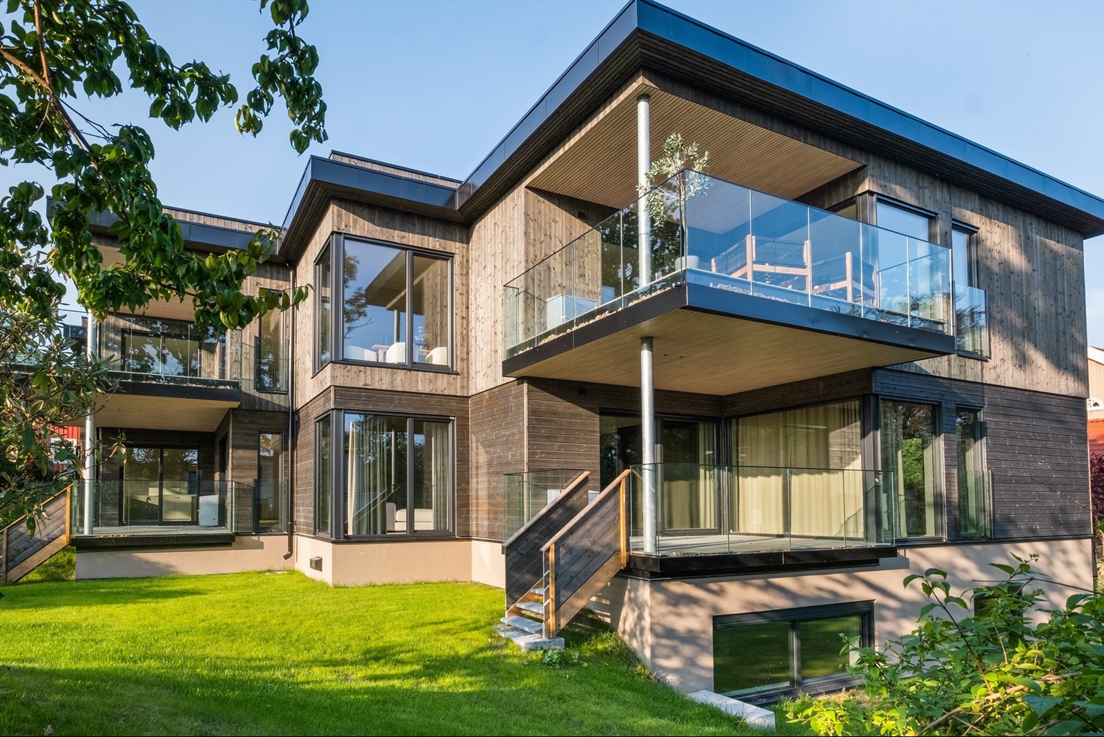 Nowoczesny dom z drewnianą elewacją i balkonami z szklanymi balustradami, otoczony zielonym trawnikiem w słoneczny dzień. Widoczne duże okna i minimalistyczny design.