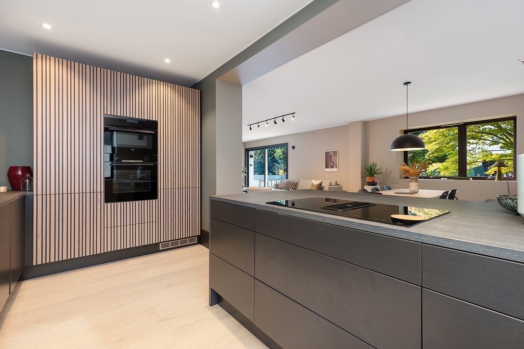 Nowoczesna kuchnia z drewnianą zabudową i wyspą kuchenną. Widok na salon przez otwarte przejście. Minimalistyczny design, naturalne światło.