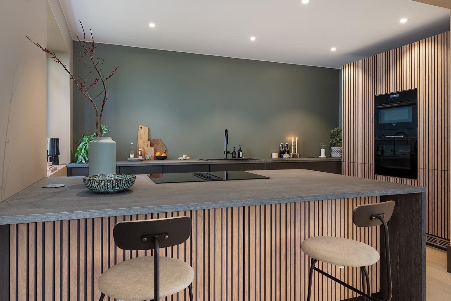 Nowoczesna kuchnia z wyspą, wykończona drewnianymi lamelami. Stonowana kolorystyka, minimalistyczny design, oświetlenie punktowe. Widoczne krzesła barowe i sprzęt AGD.