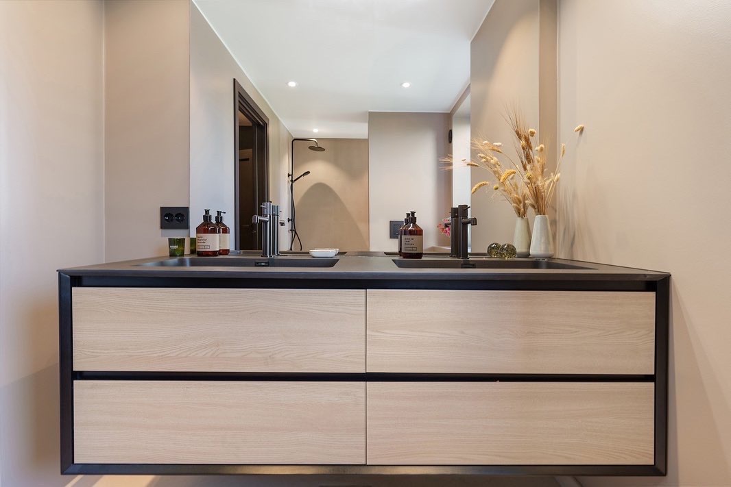 Nowoczesna łazienka z podwójną umywalką na szafce z jasnego drewna i czarnymi akcentami. W lustrze odbija się kabina prysznicowa. Minimalistyczny design.