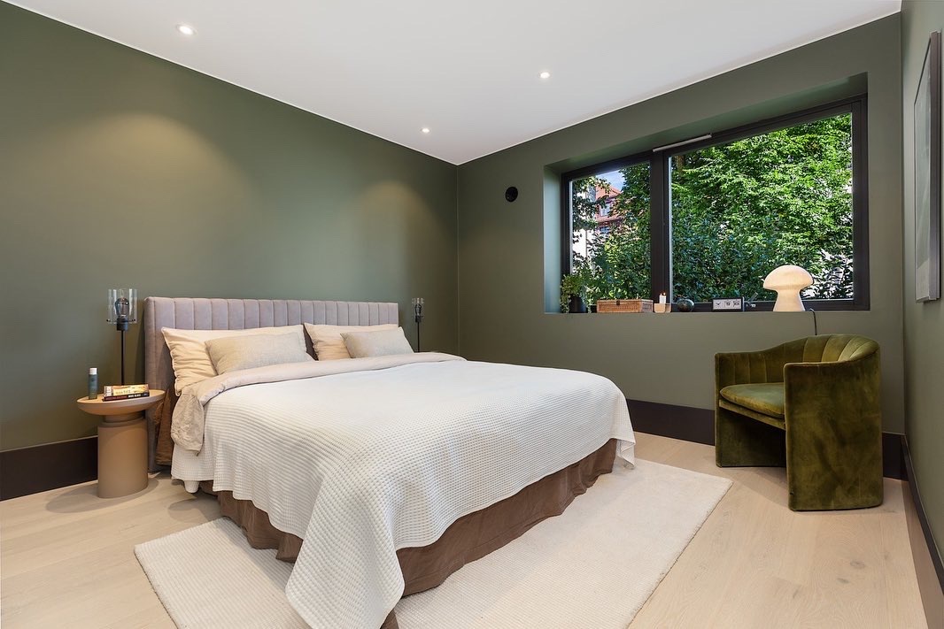 Stylowa sypialnia z oliwkowymi ścianami, jasną podłogą i dużym oknem z widokiem na zieleń. Minimalistyczny design z beżowym łóżkiem i zielonym fotelem.