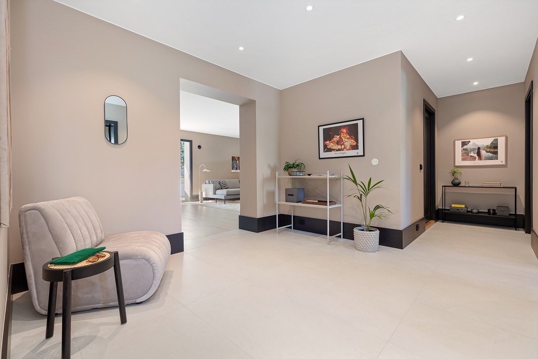 Nowoczesny hol z beżowymi ścianami, białymi płytkami podłogowymi i czarnymi listwami przypodłogowymi. Widoczne meble: fotel, stolik, regał i obrazy na ścianach.