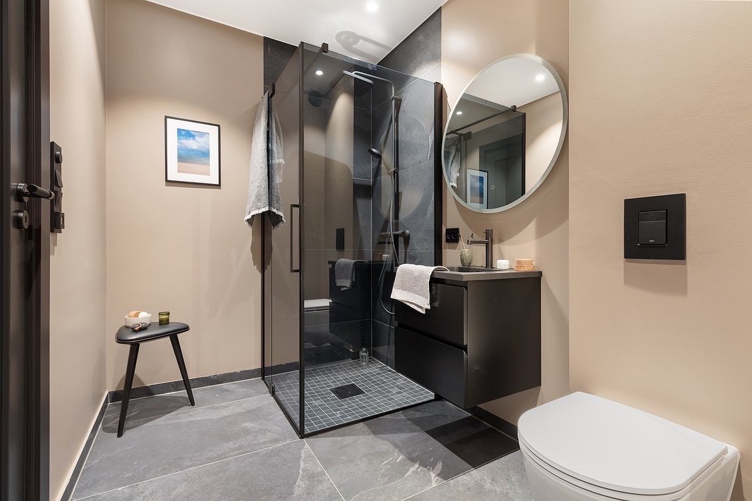 Nowoczesna łazienka z kabiną prysznicową typu walk-in, czarną armaturą i szarą płytką podłogową. Minimalistyczny design z okrągłym lustrem i beżowymi ścianami.