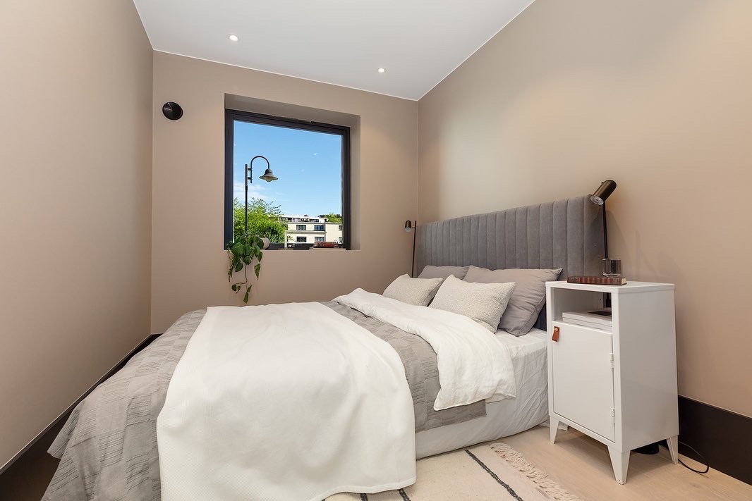 Nowoczesna sypialnia z szarą tapicerowaną ścianą za łóżkiem, białą szafką nocną i widokiem na zieleń za oknem. Minimalistyczny design, beżowe ściany, lampka nocna.