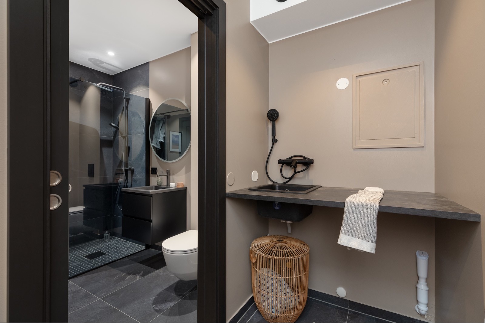 Nowoczesna łazienka z czarnymi akcentami: kabina prysznicowa, umywalka i toaleta. Beżowe ściany, rattanowy kosz i ciemna podłoga tworzą spójną całość.
