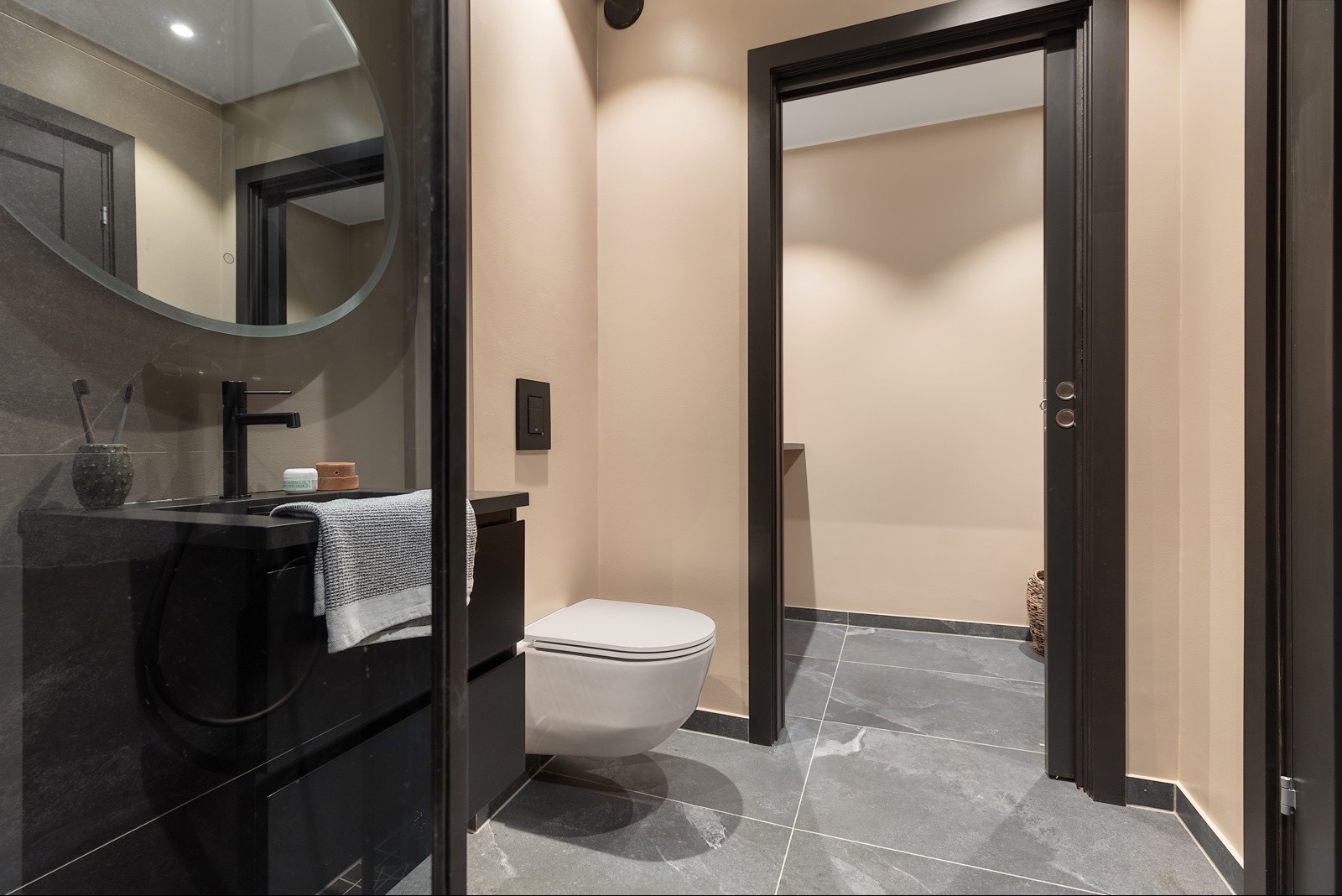 Nowoczesna łazienka z czarną umywalką, okrągłym lustrem i szarą marmurową podłogą. Beżowe ściany i ciemne ramy drzwi dodają elegancji.