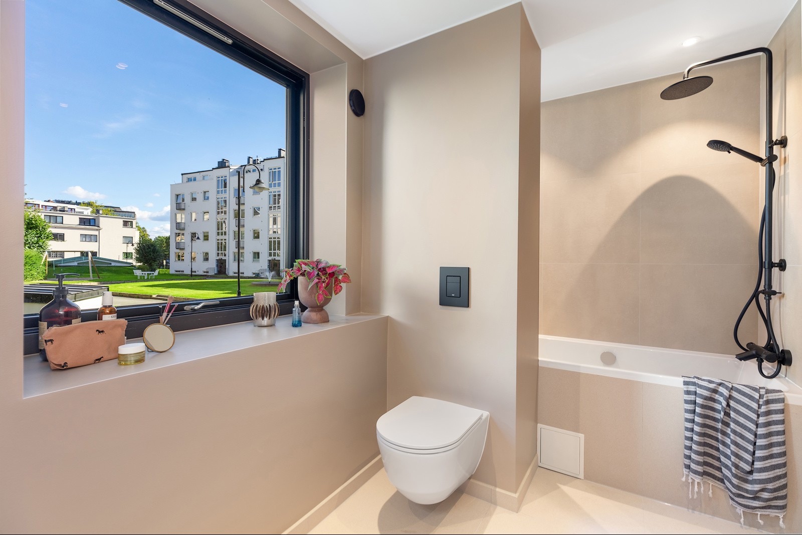 Nowoczesna łazienka w beżowych tonacjach z oknem wychodzącym na zieleń. Minimalistyczna toaleta, czarna armatura i dekoracje na parapecie okiennym.