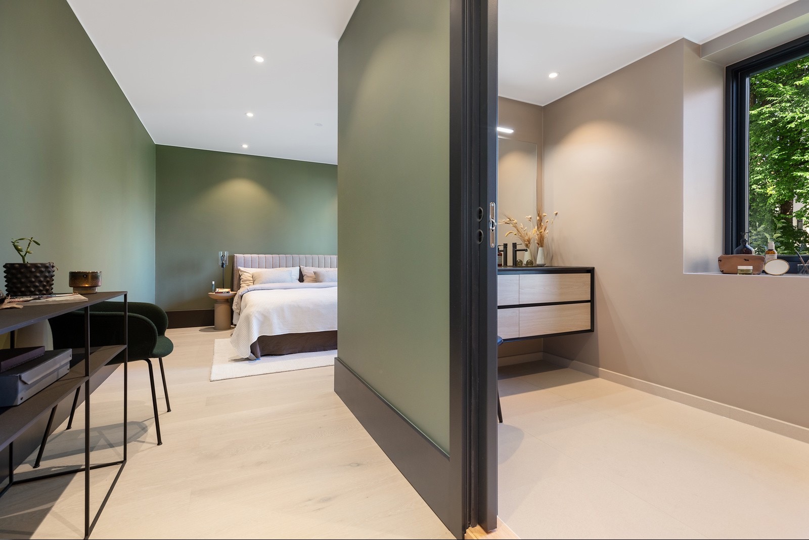 Nowoczesna sypialnia z łazienką en-suite, widok przez otwarte drzwi. Zielone ściany, drewniana podłoga, minimalistyczny design. Umywalka z szafką w łazience.