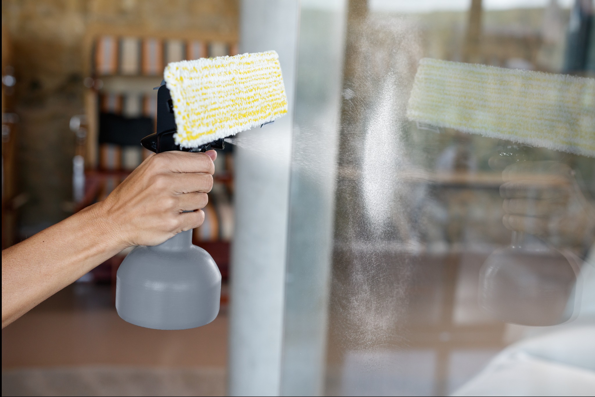 Mycie okna: ręka z rozpylaczem i myjką z żółtą nakładką, widoczne krople płynu na szybie, w tle rozmyty widok wnętrza.