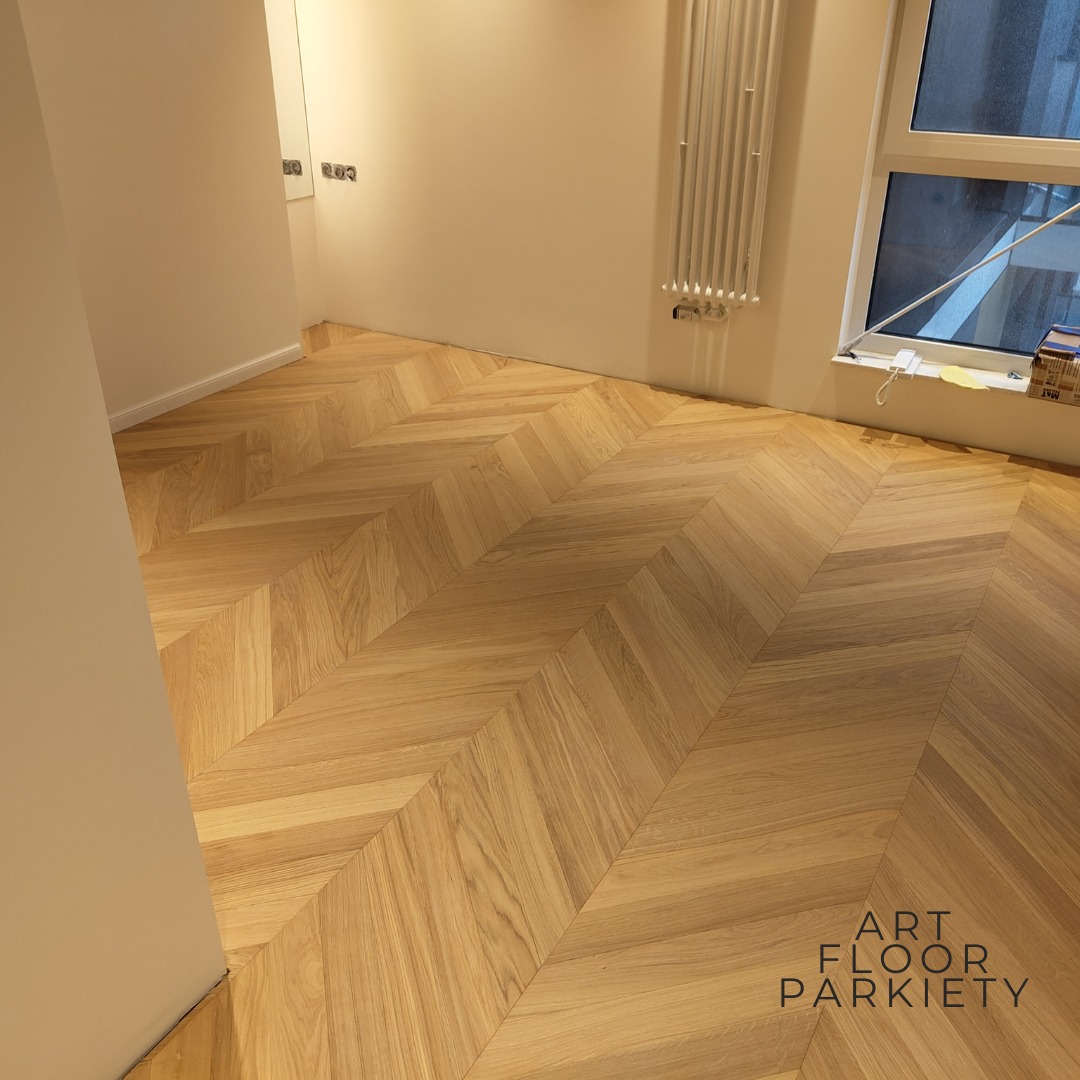 Parkiet ułożony w jodełkę w jasnym pomieszczeniu z grzejnikiem i oknem. Widoczny fragment ściany i logo firmy Art Floor Parkiety w prawym dolnym rogu.