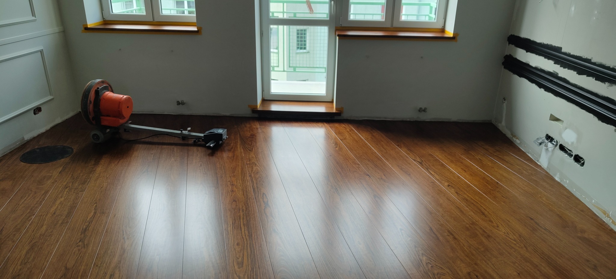 Błyszcząca, brązowa podłoga z paneli w pokoju z oknem balkonowym. Widoczna maszyna do obróbki podłóg i przygotowane ściany do malowania.