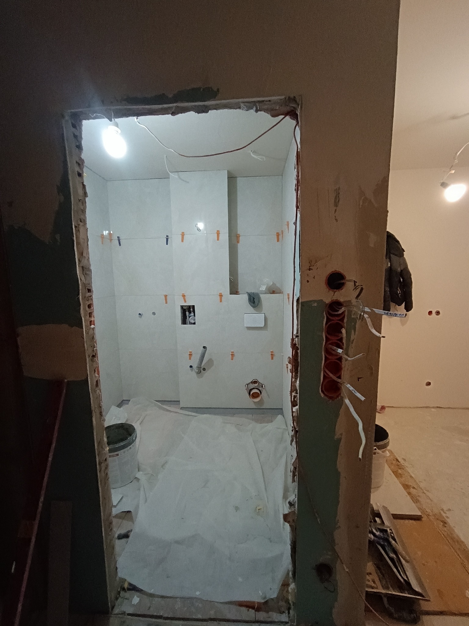 Remont łazienki w trakcie: białe płytki z krzyżykami dystansowymi, instalacja wodna i elektryczna, zabezpieczona podłoga. Widok z korytarza.