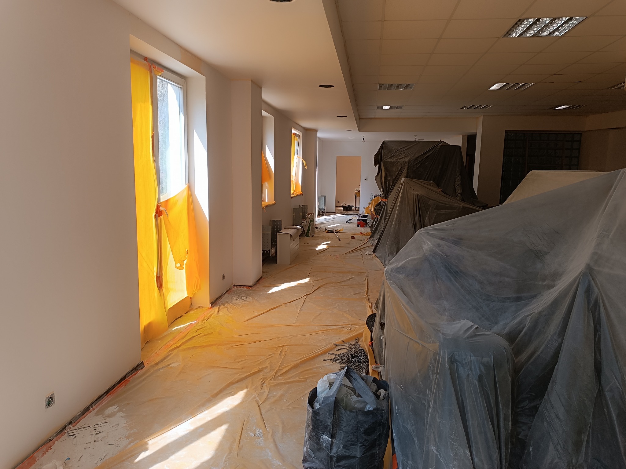 Wnętrze biura w trakcie malowania: okna zabezpieczone folią, podłoga przykryta, meble osłonięte plandekami. Widoczne ślady farby i przygotowania do prac wykończeniowych.