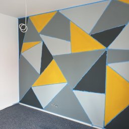 Remonovo - Ściana z geometrycznym wzorem w odcieniach szarości i żółci, zabezpieczona taśmą malarską. Widoczne gniazdka elektryczne i fragment drabiny.