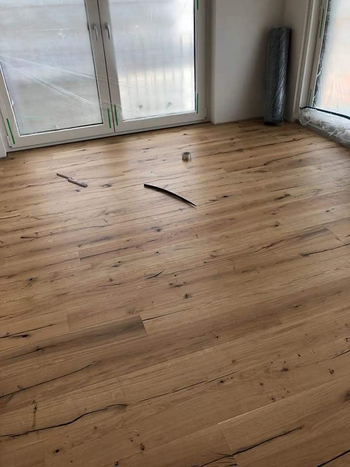 Ułożony parkiet w pokoju z oknami zabezpieczonymi folią. Widoczne narzędzia: nóż i miarka. Parkiet w odcieniach brązu z widoczną strukturą drewna.