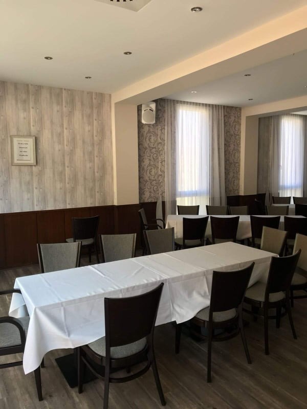 Jasne wnętrze sali konferencyjnej z drewnianą podłogą, stołami nakrytymi białymi obrusami i krzesłami. Ściany w odcieniach szarości i beżu, okna z zasłonami.