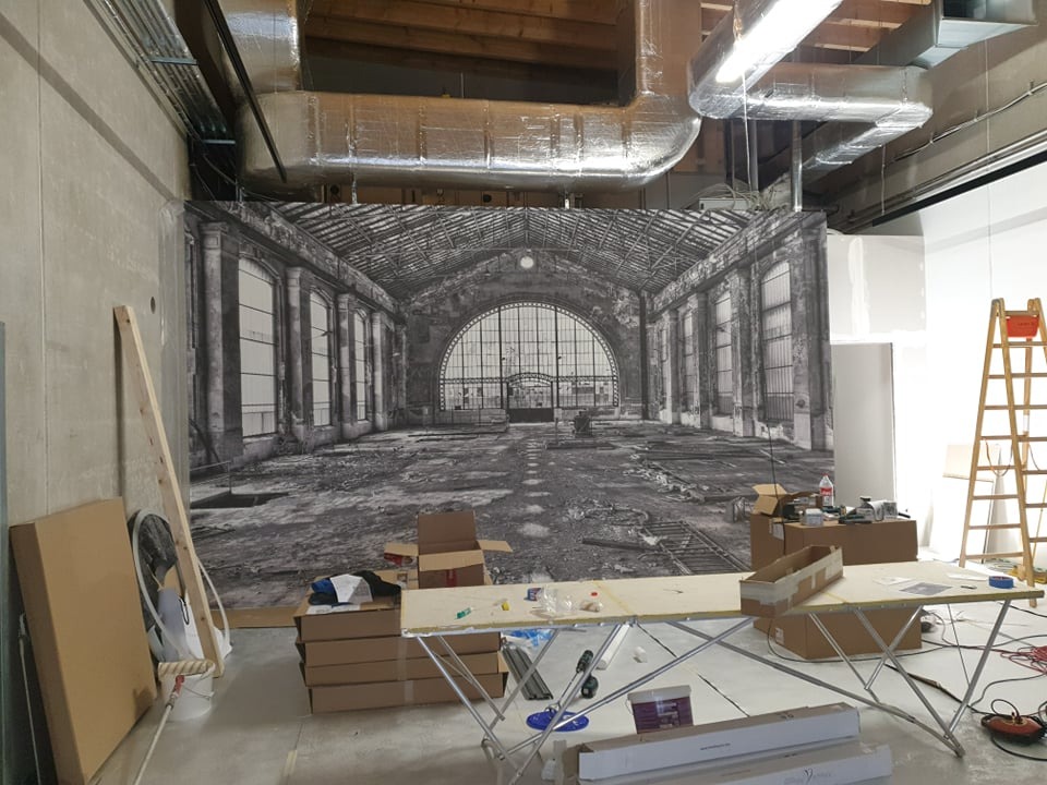 Montaż fototapety z motywem hali dworcowej w surowym wnętrzu. Widoczne narzędzia, drabina i kartony. Prace wykończeniowe w toku, surowy beton i instalacje pod sufitem.