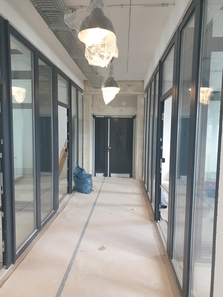 Korytarz biurowy w trakcie remontu: surowy beton, szklane ścianki działowe z czarnymi ramami, lampy zabezpieczone folią, niebieski worek na gruz. Widok w perspektywie.