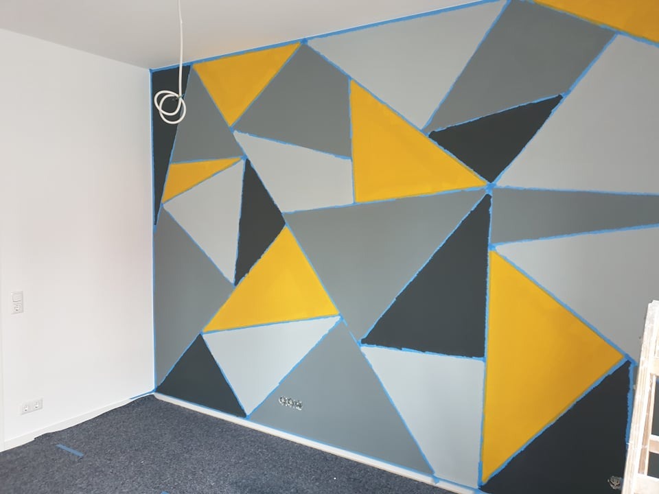 Ściana z geometrycznym wzorem w odcieniach szarości i żółci, zabezpieczona taśmą malarską. Widoczne gniazdka elektryczne i fragment drabiny.